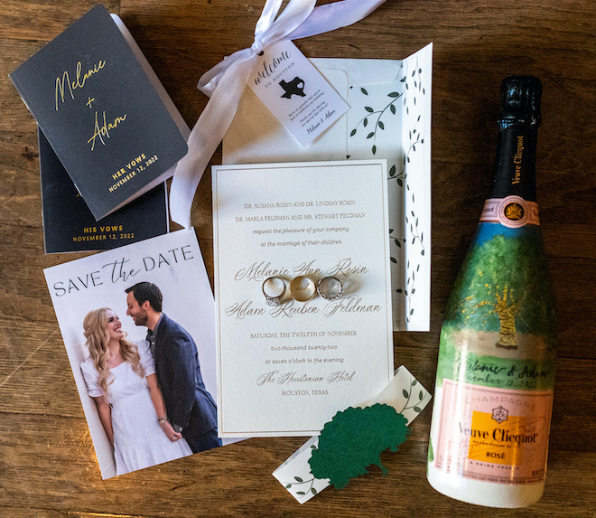 An white and gold invitation suite with a save the date, his and her vow books and a bottle of rosé.