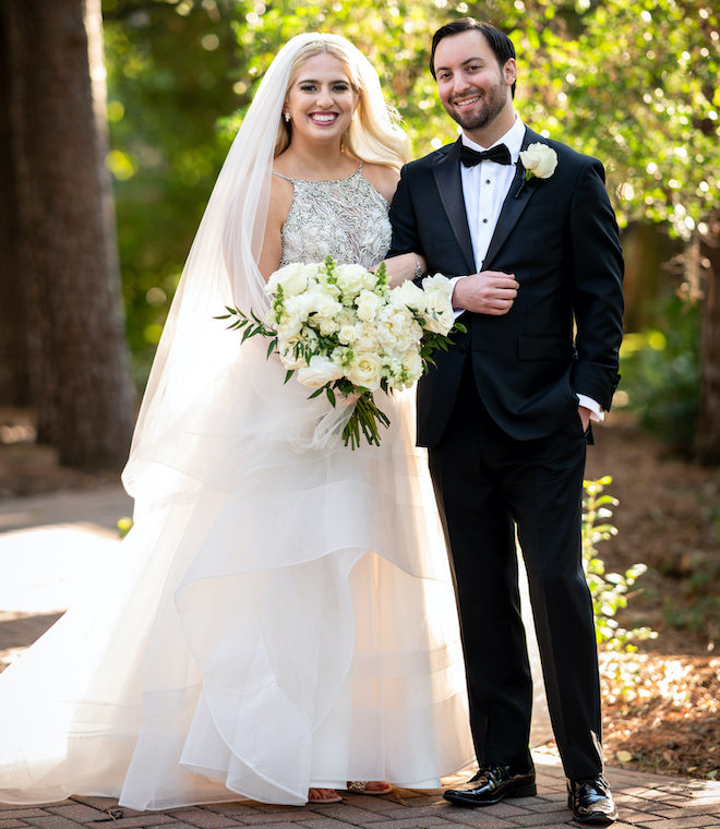 The bride and groom smiling outside. The bride is holding a bouquet with white florals and greenery. 