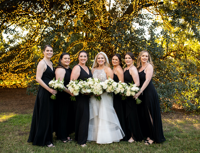 The bride and her bridesmaids in black dresses holding white and green bouquets smiling in front of the lit-up oak tree. 