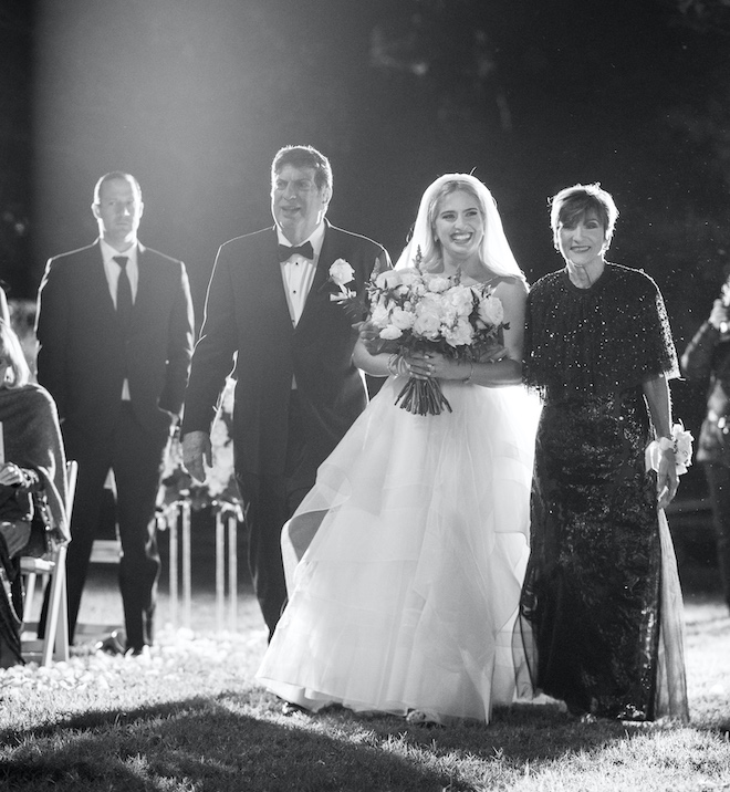 The bride walking down the aisle with her parents at nighttime. 