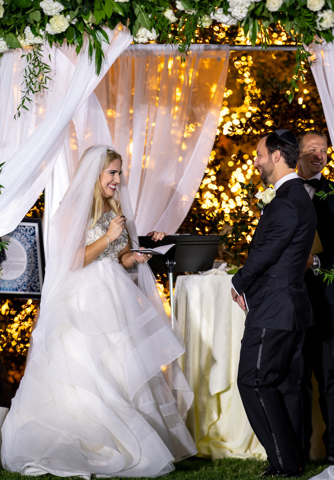 The bride smiling at the groom during their twilight wedding ceremony in front of a lit oak tree. 