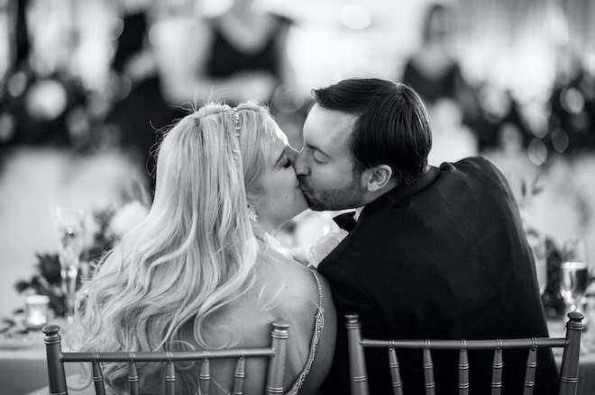 The bride and groom kissing while sitting at the reception table. 