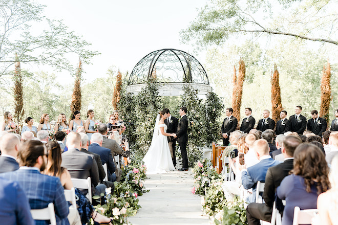 The bride and groom hold hands at the altar while exchanging vows at their outdoor ceremony. 
