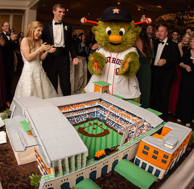 A bride and groom clapping with the Astros mascot admiring their cake replica of the Astros stadium. 