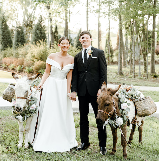 The bride and groom smile as they pose beside two donkeys at their wedding cocktail hour. 