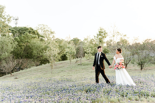 The bride and groom hold hands as they walk through a field of bluebonnets at sunset.