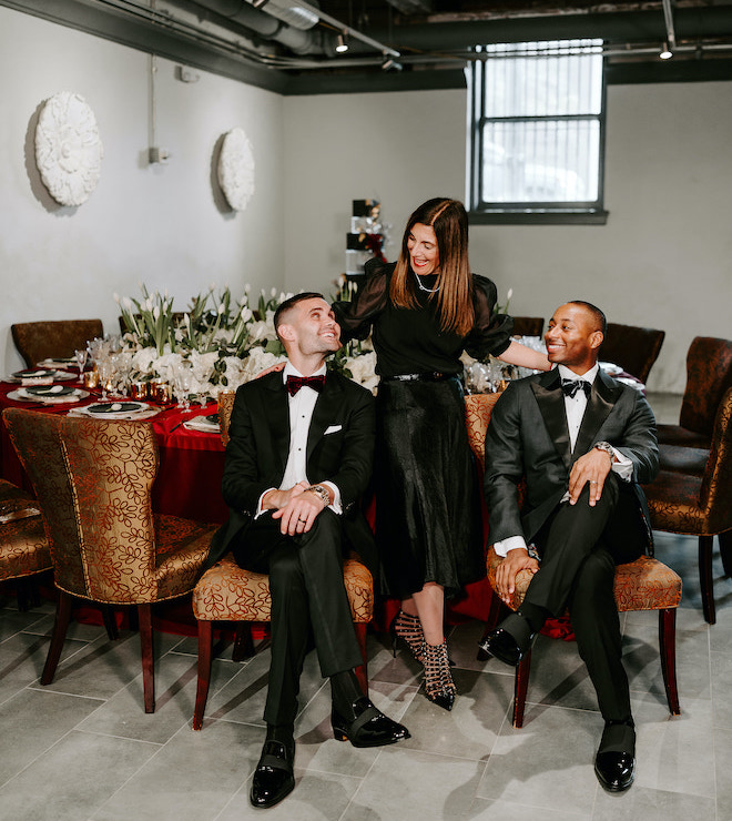 Two grooms sitting at a reception table looking up and smiling at the wedding planner, Marcela Bogado.