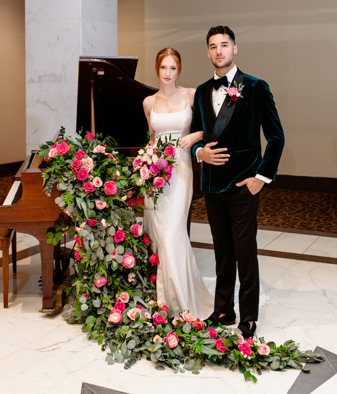 The bride and groom posing in front of a piano covered in greenery and pink florals.