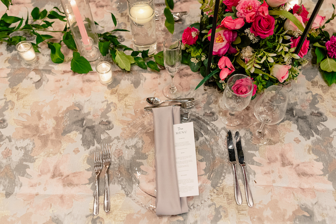A gray napkin and menu resting on a clear glass plate on the pink and gray reception table. 