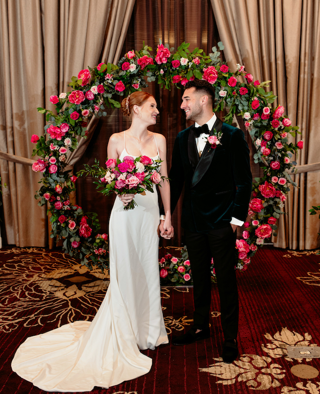 The bride and groom holding hands smiling at each other in front of the circular altar fill of greenery and pink flowers. 
