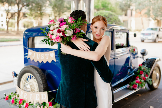 The bride hugging the groom in front of the blue vintage car.
