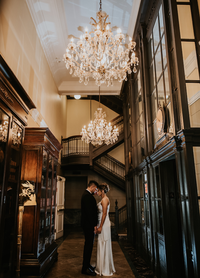 A couple holding hands in Hotel Icon under chandeliers and a staircase. 