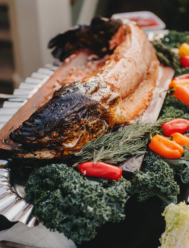 A fish cooked by Bailey Connor Catering is served to wedding guests on a wooden platter with kale and an assortment of bell peppers. 