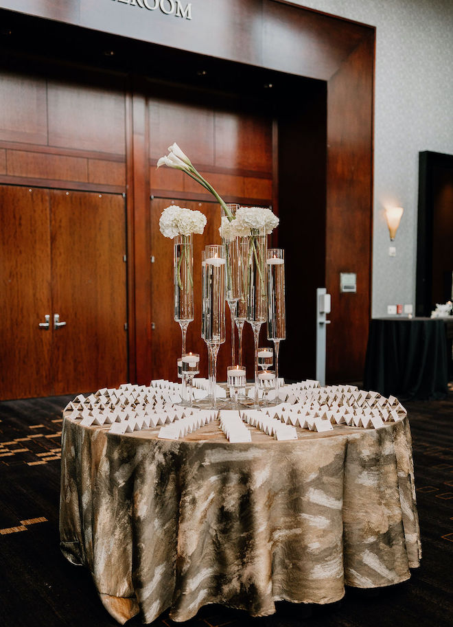 A round table with gold linens holding place cards for the guests with white floral centerpieces sitting outside the ballroom. 