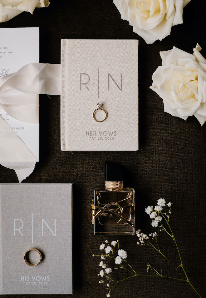 A flatlay of beige and gray books for the couple's vows, a gold perfume bottle, an engagement ring and white florals on a black background. 
