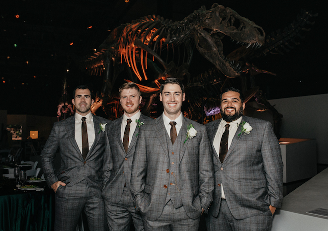 The groom and his groomsmen smile under a T-Rex fossil at the wedding reception. 