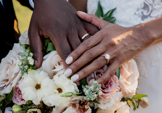 The bride and groom place their hands on top of the pink and white wedding bouquet to show off their wedding rings.