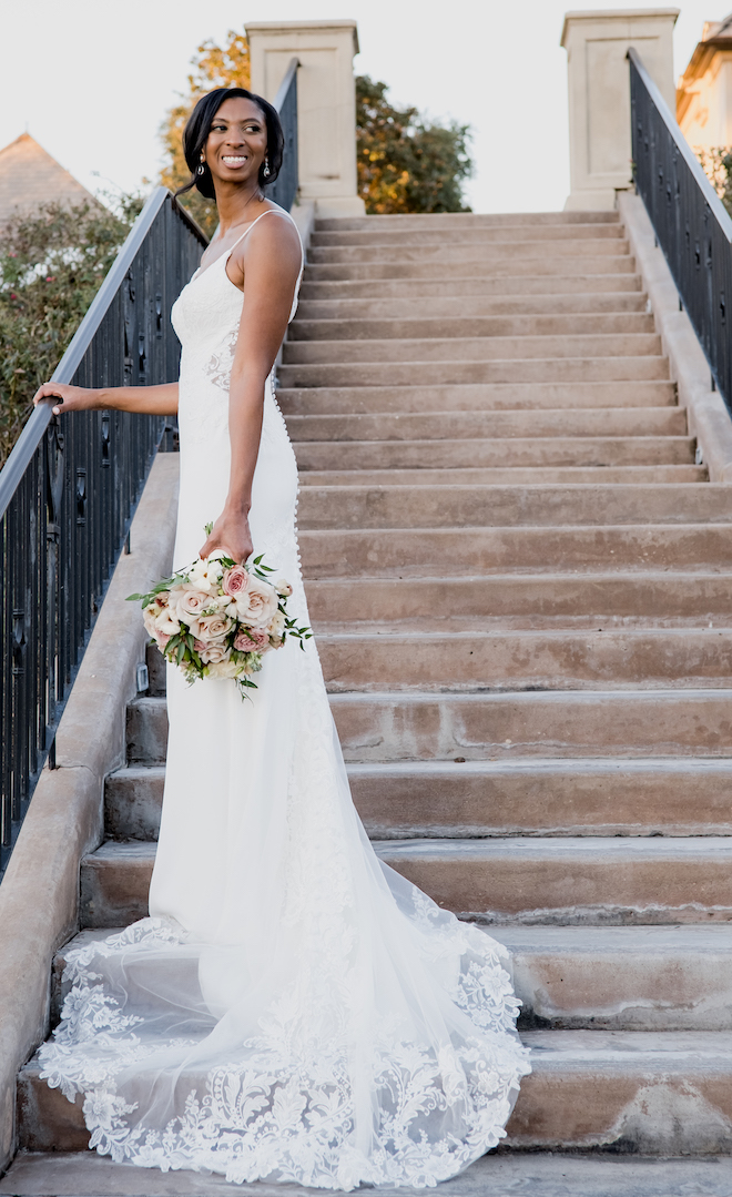 The bride smiles while holding her pink and white wedding bouquet on the stairs outside her wedding venue. 
