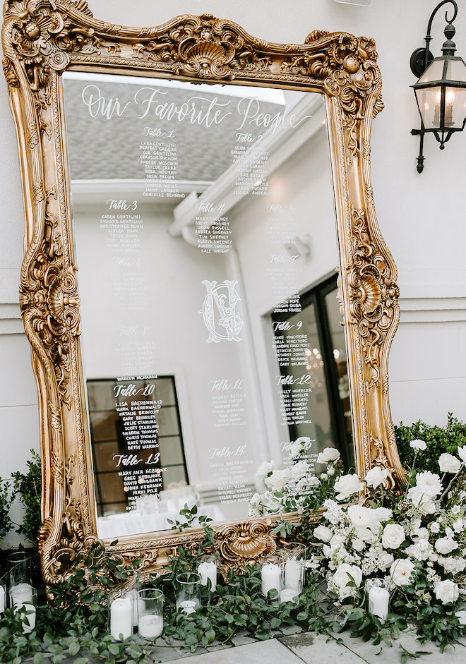 A gold mirrored seating chart with greenery and white florals at the bottom. 