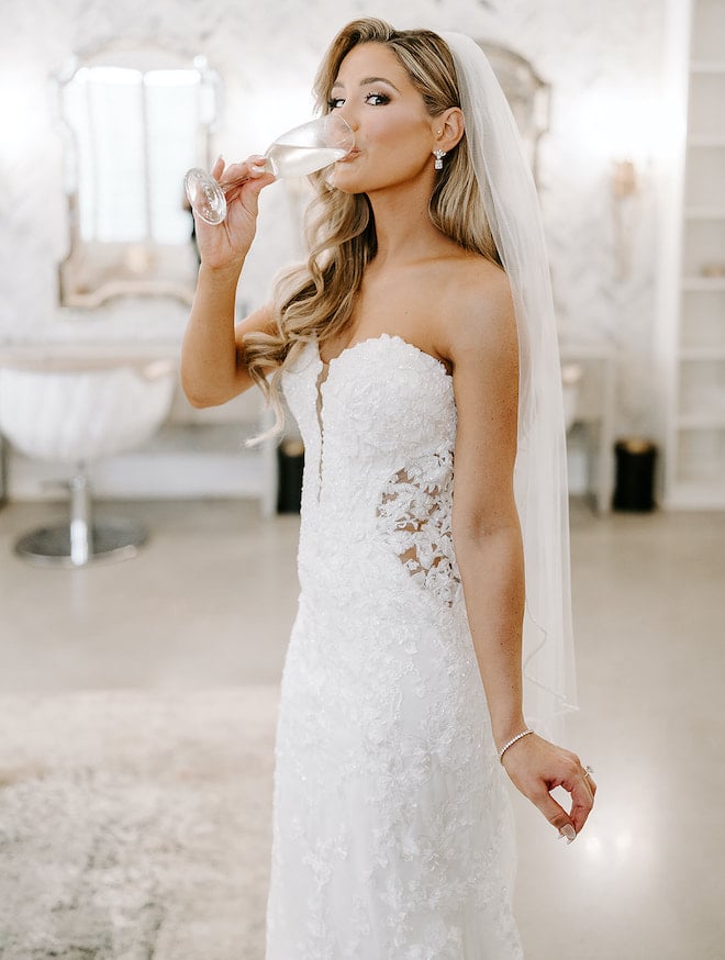 The bride sipping champagne in a strapless lace wedding gown and veil. 