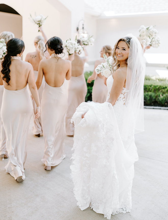 The bride smiling while her bridesmaids walk in front of her in pink satin gowns with white bouquets. 
