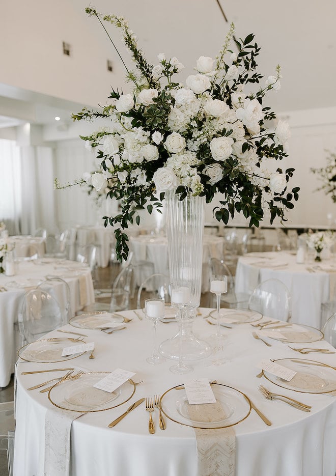 A white reception table with a tall white floral and greenery centerpiece. 