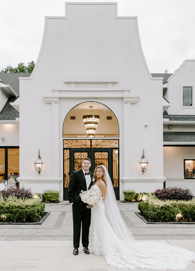 The bride and groom smiling in front of their white wedding venue. 