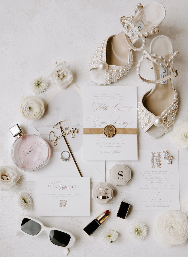 A white and gold flatlay with Jimmy Choos, white florals, white glasses and a Chanel perfume bottle. 