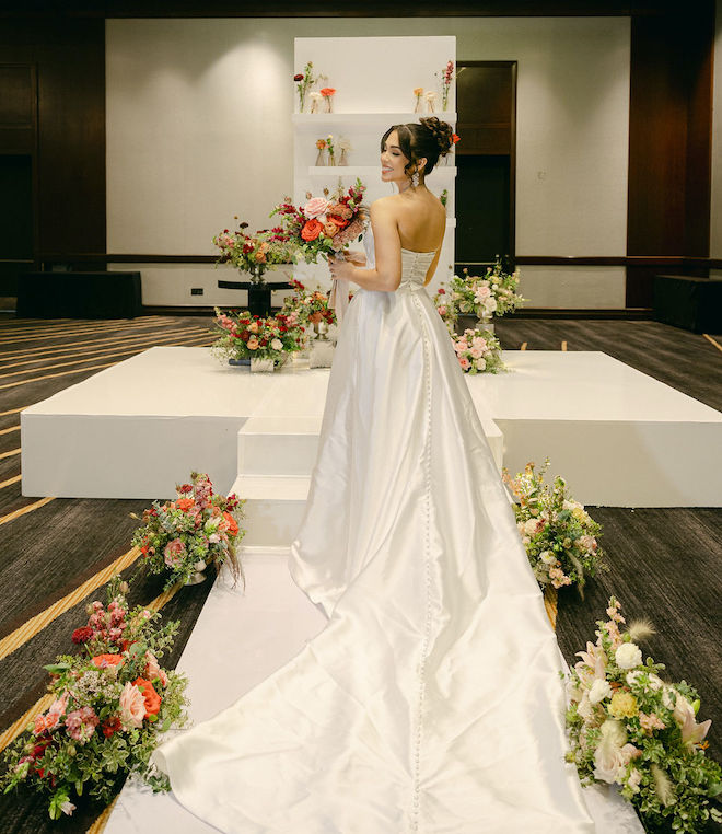 The bride looks over her shoulder and smiles as she walks down the aisle surrounded by florals at the Royal Sonesta Houston Galleria.