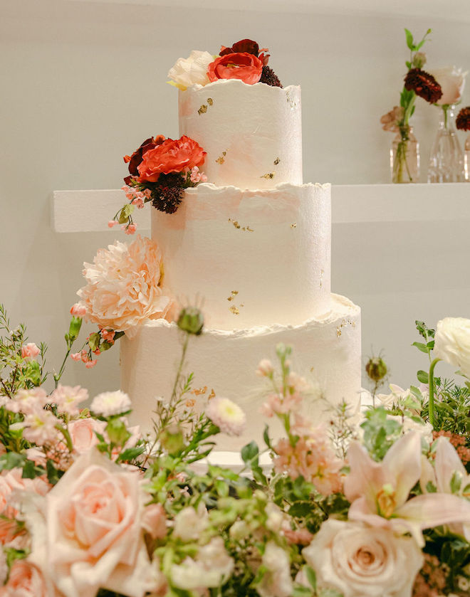 A three-tier wedding cake embellished with florals created by Susie's Cakes is surround by florals at the wedding reception.