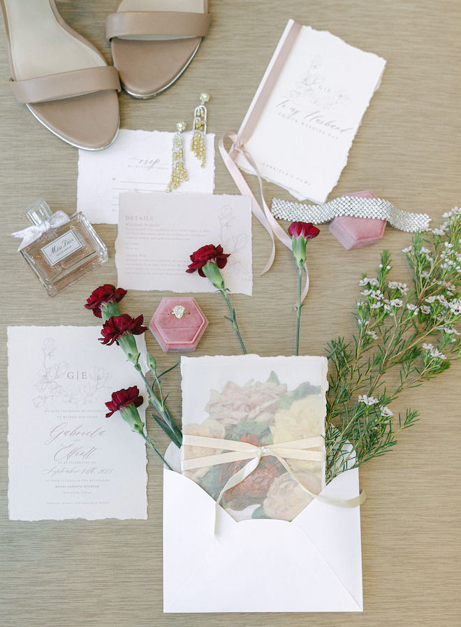 A flatlay of the wedding invitations and stationery, florals, jewelry, perfume and bridal shoes. 