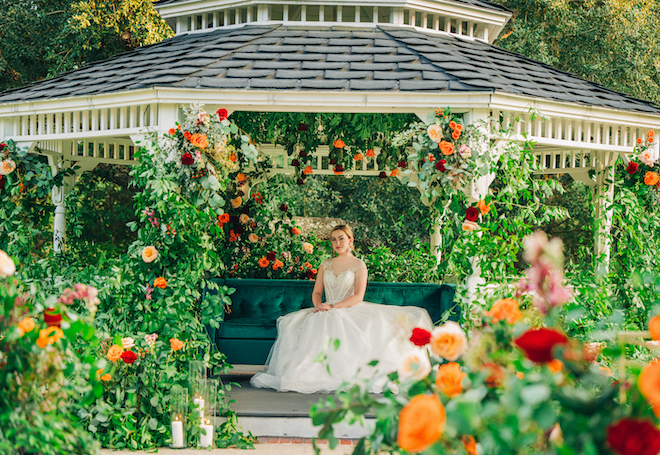The bride poses on the emerald green couch under the garden gazebo decorated in autumn-colored flowers at her outdoor ceremony.