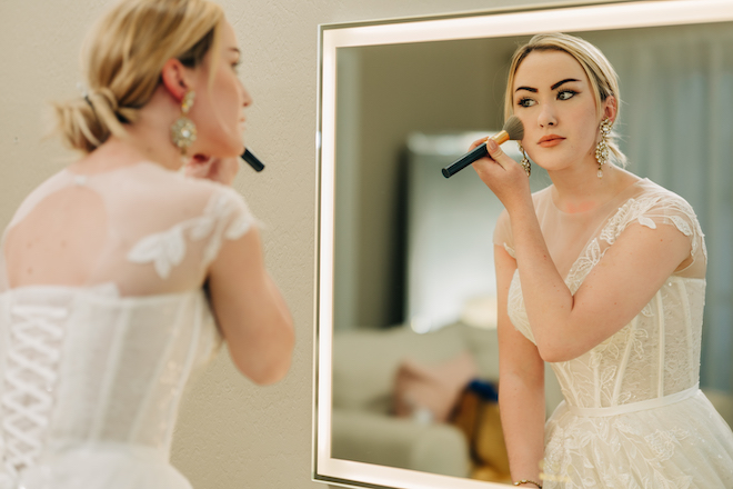 A bride looking in the mirror while putting on blush. 