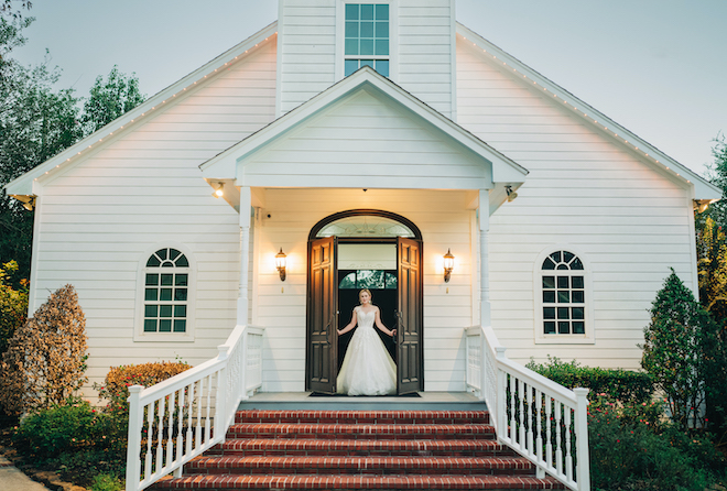 The bride walks out of the doors of the white chapel to go outside for her wedding ceremony. 