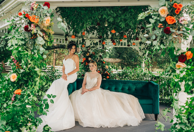 The two brides sit on an emerald green sofa under the garden gazebo decorated in flowers. 