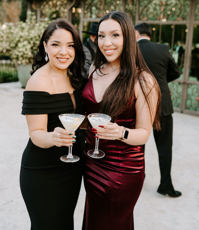 Two women wearing black tie gowns holding cocktails. 