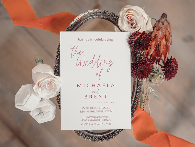 Orange and white wedding stationery lay on an antique mirror surrounded by burgundy, orange and white flowers. 