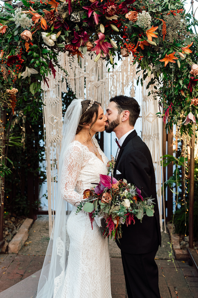 The bride and groom kiss under a floral arch at their bohemian wedding. 