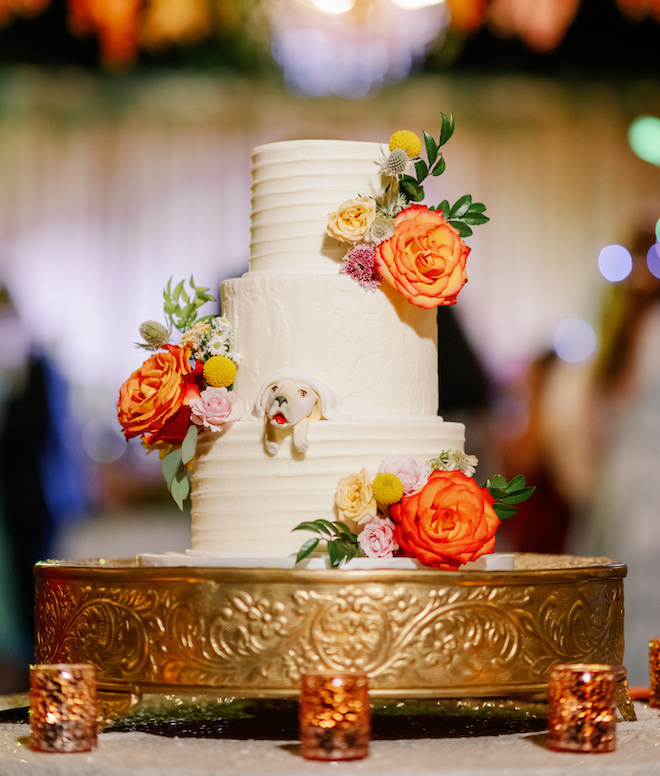 A three-tier white cake with a replica of their dog peaking out and orange florals on each tier. 