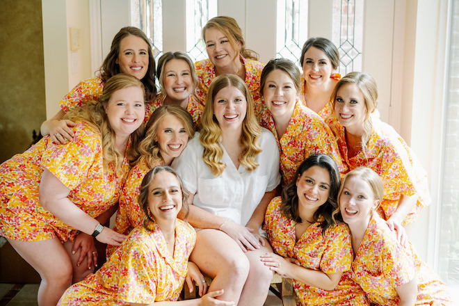 The bride smiling with her bridesmaids wearing orange and pink floral pajamas. 