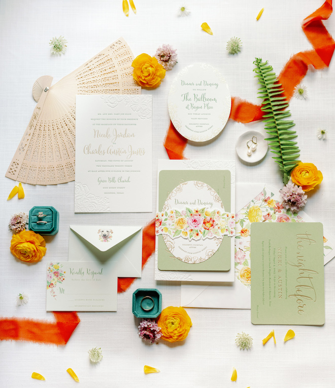 Colorful flatlay with a white and green invitation suite, a fan, wedding rings and florals. 