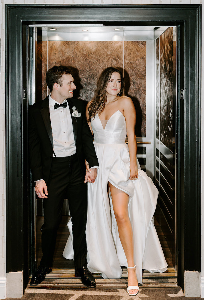 The bride and groom step out of the elevator holding hands for their city-chic ballroom wedding. 