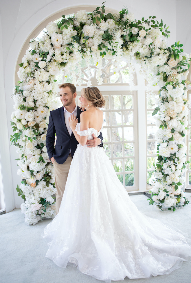 Bride with updo wearing a off-the-should wedding dress hugs a groom aside a floral installation at a Houston wedding venue. 