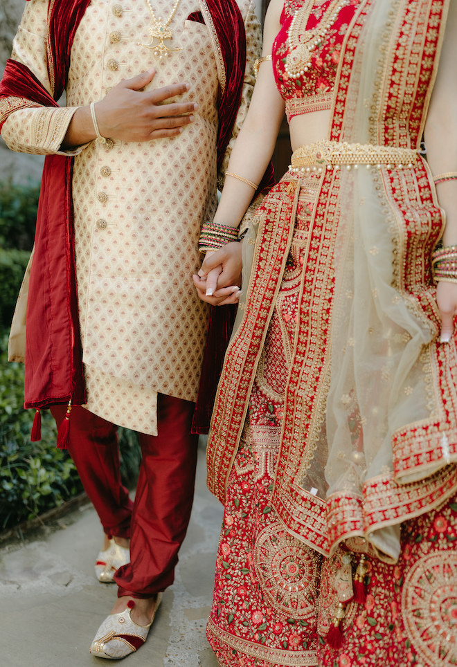 The bride and groom hold hands in their red and gold traditional Indian wedding attire.