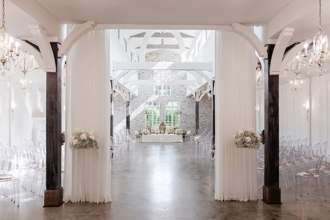 The ceremony space with clear chairs, chandeliers and floral arrangements. 