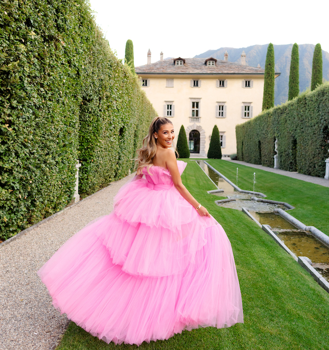 A girl smiling looking back in a pink dress as she runs on a field of grass toward an Italian building. 