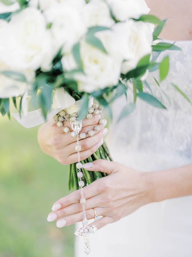 A silver colored rosary wraps around the bride's wedding bouquet of white roses.