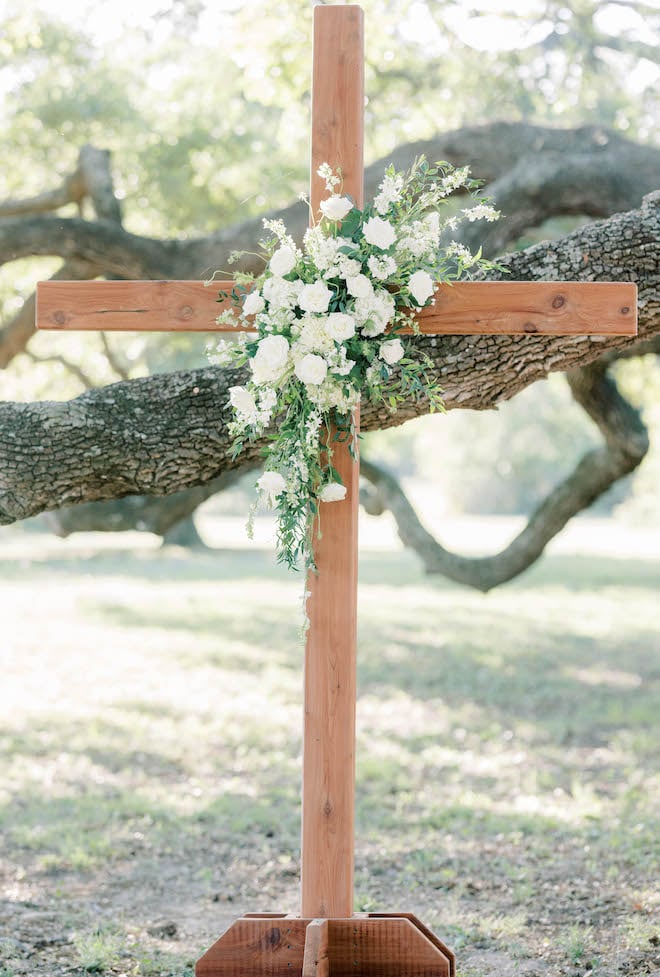 A wooden cross with white roses and greenery is placed under an oak tree for the outdoor wedding ceremony.