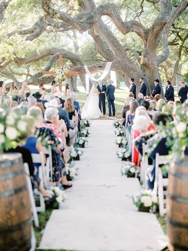 The bride and groom exchange vows under an oak tree on their family land in Brenham, Texas for a sentimental Texas ranch wedding.