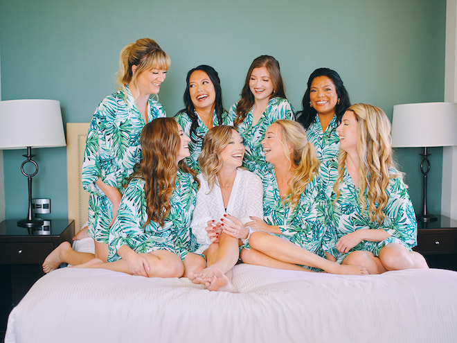 A bride in white and her bridesmaids in tropical robes laughing on a bed. 